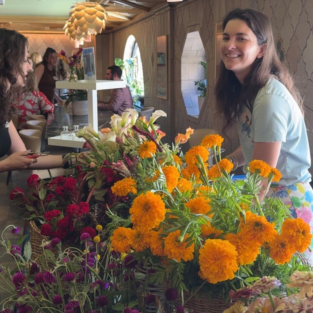 A smiling woman stands behind a large, vibrant display of fresh local flowers, including prominent orange marigolds, red celosia, and purple blossoms, inside the S&W building.
