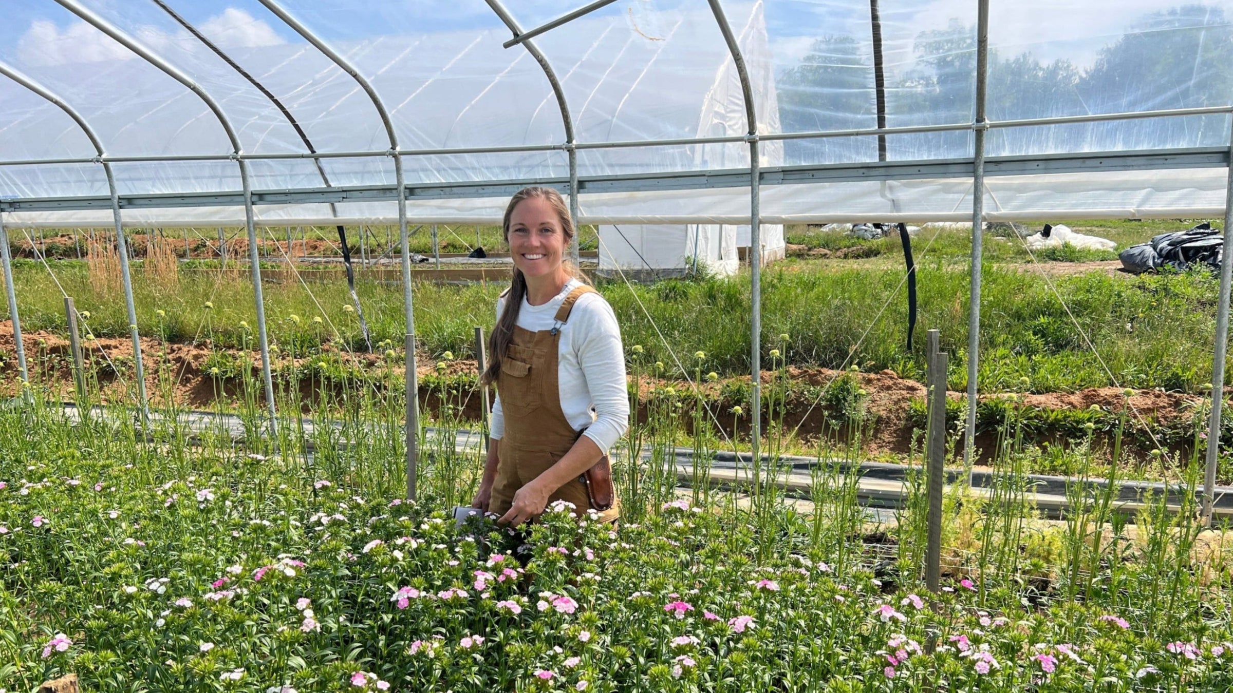A smiling woman stands in a greenhouse among rows of pink and white flowers, wearing a brown apron over a white long-sleeve shirt.